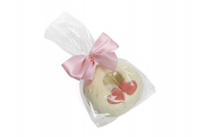 Ein weißer Baumkuchenring verziert mit zwei rosafarbenen Herzen, eingepackt in eine Folietüte und mit einer rosafarbenen Schleife zugebunden. Der Link zeigt das Bild in groß.