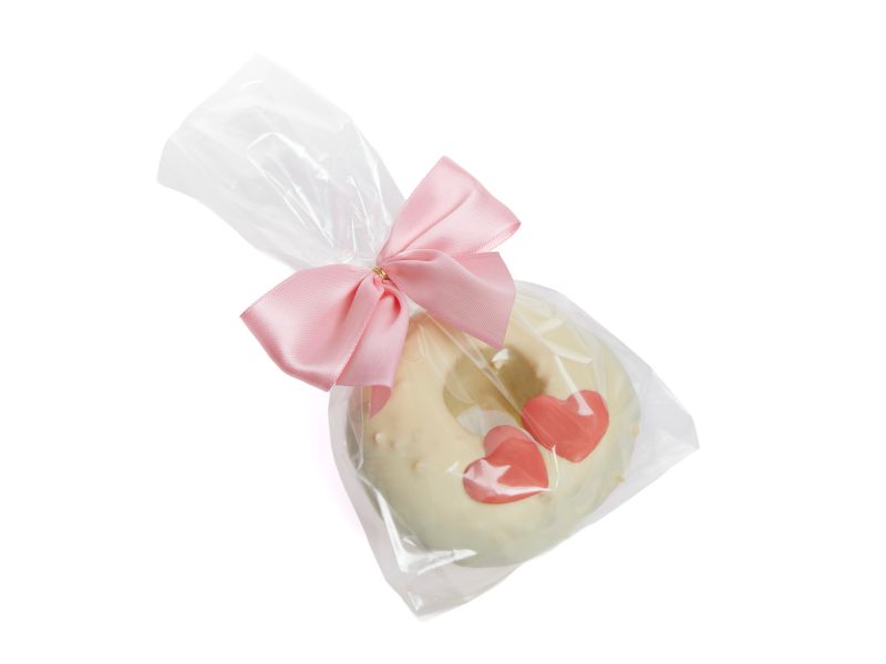 Ein kleiner weißer Baumkuchenring mit zwei rosanen Herzen drauf in einer transparenten Geschenktüte eingepackt.