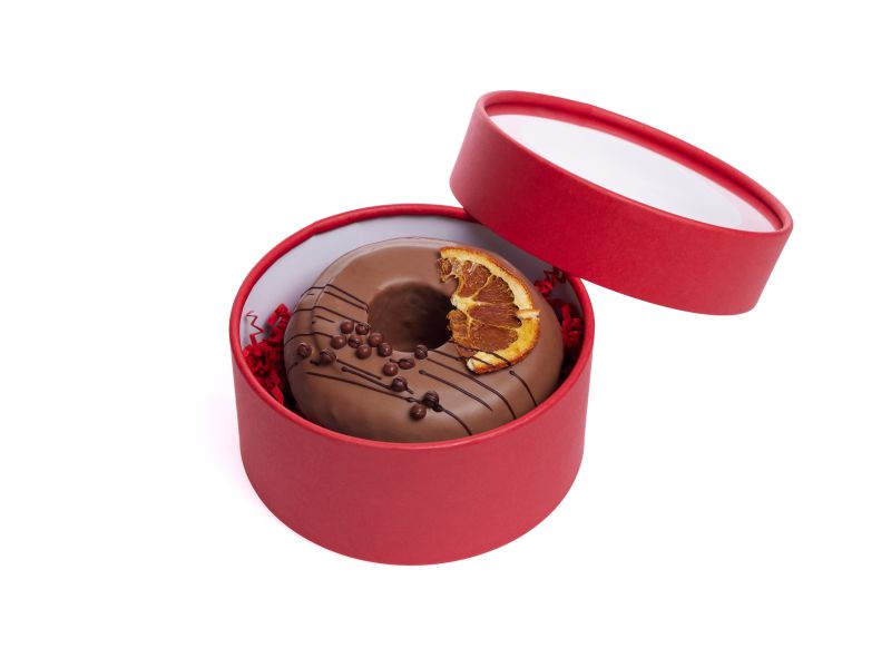 Ein mit vollmilch überzogener Baumkuchenring ist mit einer Orangenscheibe und zartbitter Schokoladenstreifen dekoriert und liegt in einer bordeauxfarbenen Präsentedose mit Deckel.